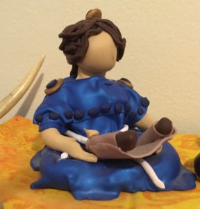 Polymer clay statue of a woman with deeper beige skin, dark brown hair, and a blue dress, reading a scroll. 