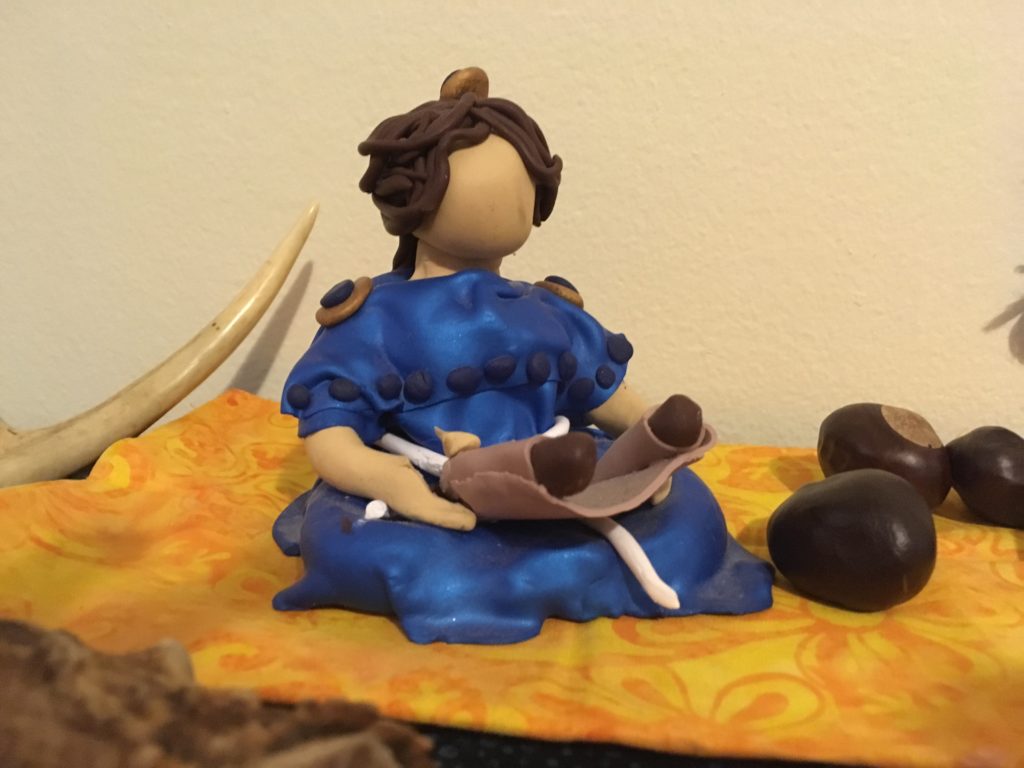 Photograph of polymer clay figure of a woman reading a scroll, with several horse chestnuts beside her, and the tip of an antler in the background
