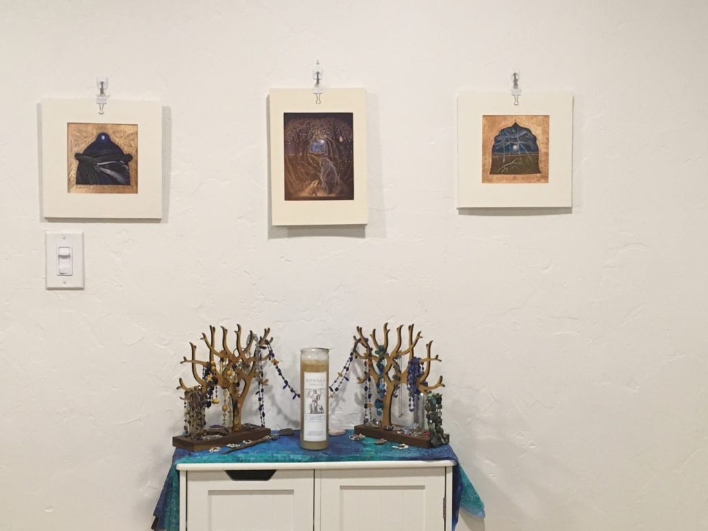 Venus and Jupiter shrine: two wooden trees holding necklaces and jewellery on a small set of shelves with a large glass-enclosed candle in the middle and a blue-green silk altar cloth. Three prints hang above.