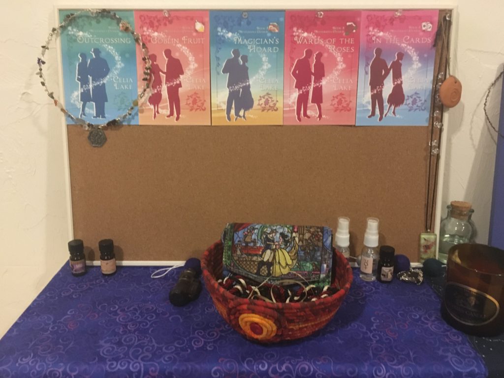 Writing shrine with corkboard with book covers pinned to it, along with a few necklaces. A red cloth bowl sits in the center on a blue cloth, with a wallet with a stained glass image from Beauty and the Beast.. There are several small perfume and spray bottles.
