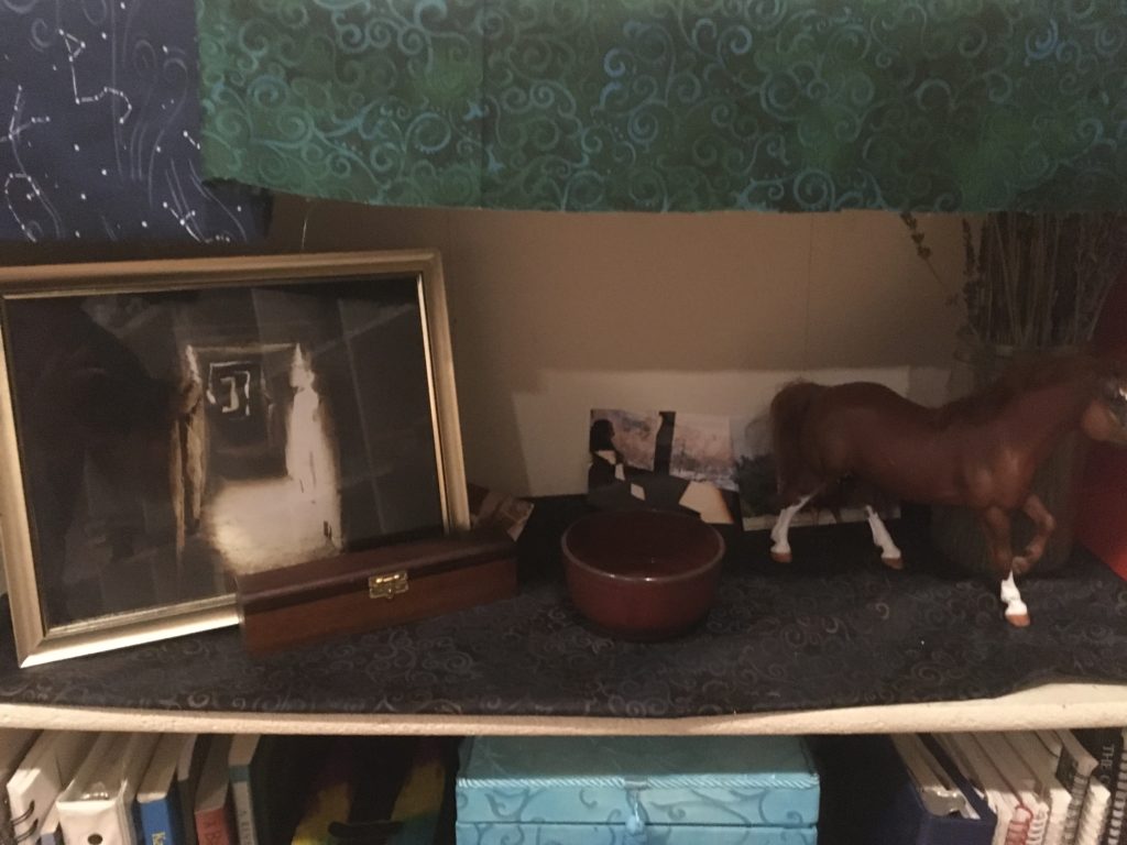 Ancestor shrine, with a large framed image of a barrow, a wooden box that is long and narrow, a brown ceramic bowl, and a model of a chestnut horse with white legs, as well as some photographs. 