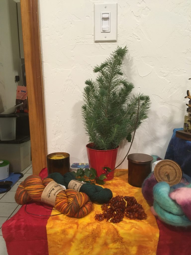 Shrine decorated for yuletide, with a potted small pine tree, two candles in amber glass jars, two skeins of yarn (yellow-browns and green), some wool waiting to be spun, and a pile of amber beads on a dark red altar cloth with a golden yellow runner. Further details in the text.