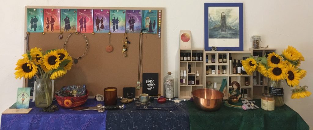 A view of my shrine, as of August 2020: bookshelves with altar cloths of blue, black, and green dividing the space, a bulletin board with cover images of books and jewellery hanging from it, various bowls for offerings and ongoing workings, wooden shelving holding bottles of oils and perfumes. Two bright golden vases of sunflowers are at either end. 